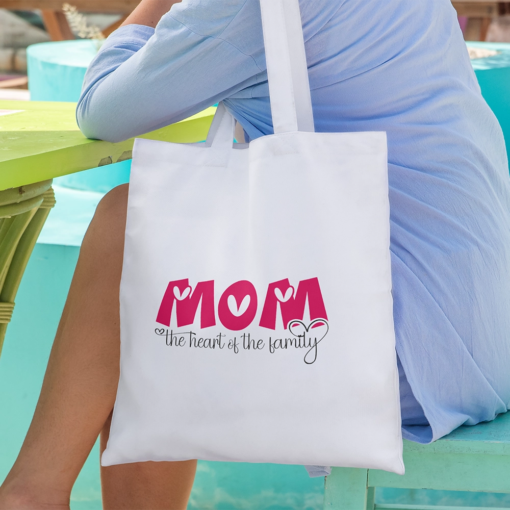 Tote bag bianca dedicata alla mamma con scritta Mom The Heart of the Family, indossata in ambientazione estiva.