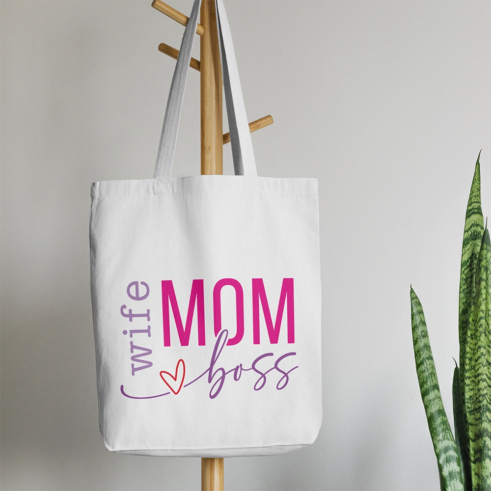 Tote bag bianca dedicata alla mamma con scritta Wife Mom Boss in lilla e rosa, appesa accanto a pianta verde.
