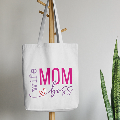 Tote bag bianca dedicata alla mamma con scritta Wife Mom Boss in lilla e rosa, appesa accanto a pianta verde.