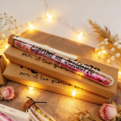 Ampollina dei Desideri con candelina rosa e paiettes, appoggiata su scatoline kraft Piccola Magia in ambientazione calda con lucine