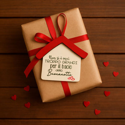 Pacchetto regalo in carta kraft con nastro rosso e casetta in legno con frase “Non si è mai troppo grandi per il bacio della buonanotte”