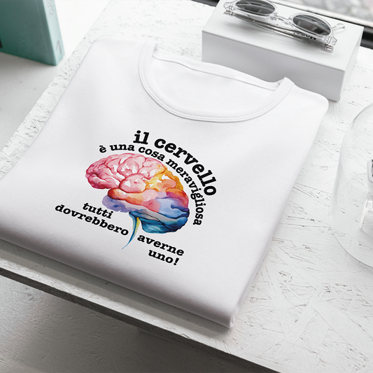 La T-shirt bianca Cervello de Il mio negozio, con un cervello colorato e la frase "Il cervello è una cosa meravigliosa, tutti dovrebbero averne uno!", è appoggiata vicino agli occhiali da sole su una superficie strutturata: una divertente maglietta personalizzata!.