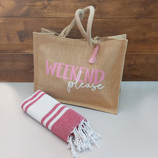 Il Combo Borsa Juta e Telo Fouta WEEKEND di In Casa di Ale è composto da una borsa in juta color tan con la scritta "WEEKEND please" in rosa/bianco e da un asciugamano fouta a righe rosse, ideale per le fughe estive del 2025.