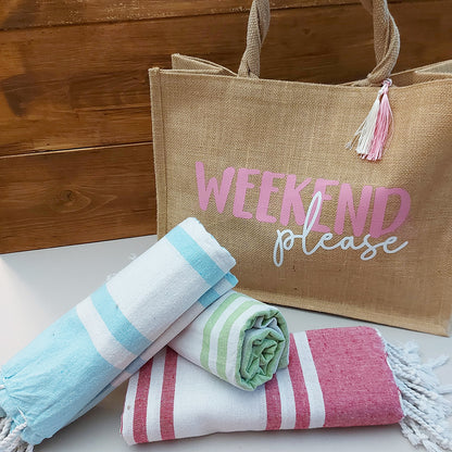 Il Combo Borsa Juta e Telo Fouta WEEKEND di In Casa di Ale è composto da una borsa in juta color tan con la scritta "WEEKEND please" in rosa/bianco e da un asciugamano fouta a righe rosse, ideale per le fughe estive del 2025.