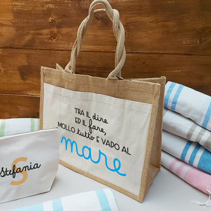 Il Combo Borsa Juta, Telo Fouta e Pochette di Casa di Ale presenta una borsa in juta con testo in italiano e scritte in blu, un sacchetto con zip Stefania bianco con una S arancione e un Telo Fouta a righe, il tutto su una superficie chiara con uno sfondo in legno.