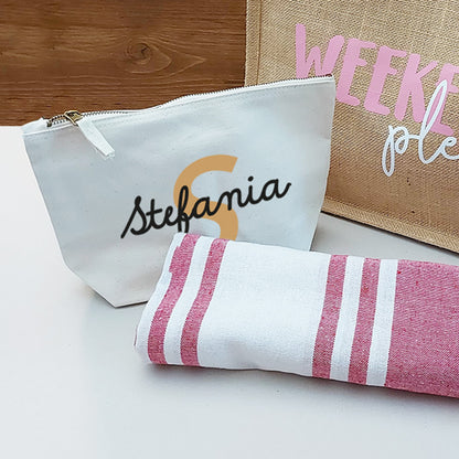 Il Combo Borsa Juta, Telo Fouta e Pochette di Casa di Ale presenta una borsa in juta con testo in italiano e scritte in blu, un sacchetto con zip Stefania bianco con una S arancione e un Telo Fouta a righe, il tutto su una superficie chiara con uno sfondo in legno.