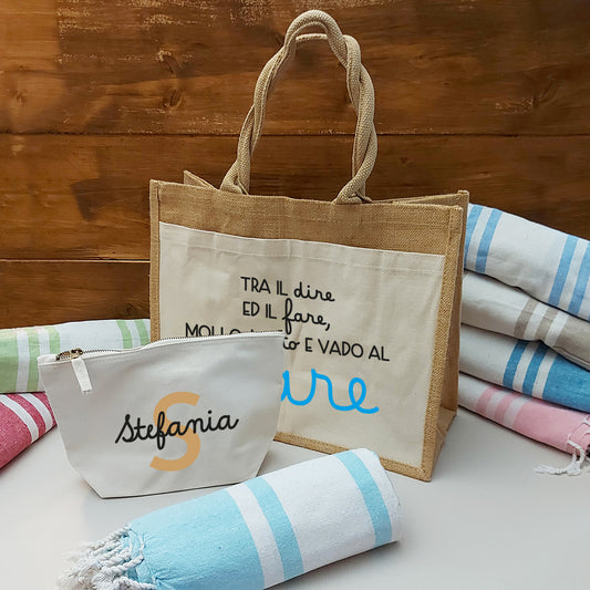 Il Combo Borsa Juta, Telo Fouta e Pochette di Casa di Ale presenta una borsa in juta con testo in italiano e scritte in blu, un sacchetto con zip Stefania bianco con una S arancione e un Telo Fouta a righe, il tutto su una superficie chiara con uno sfondo in legno.