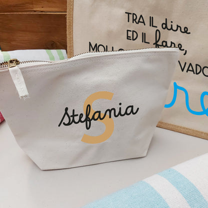 Il Combo Borsa Juta, Telo Fouta e Pochette di Casa di Ale presenta una borsa in juta con testo in italiano e scritte in blu, un sacchetto con zip Stefania bianco con una S arancione e un Telo Fouta a righe, il tutto su una superficie chiara con uno sfondo in legno.
