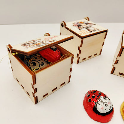Scatolina pasquale in legno aperta con paglina verde e cioccolatini a forma di ape e coccinella all’interno