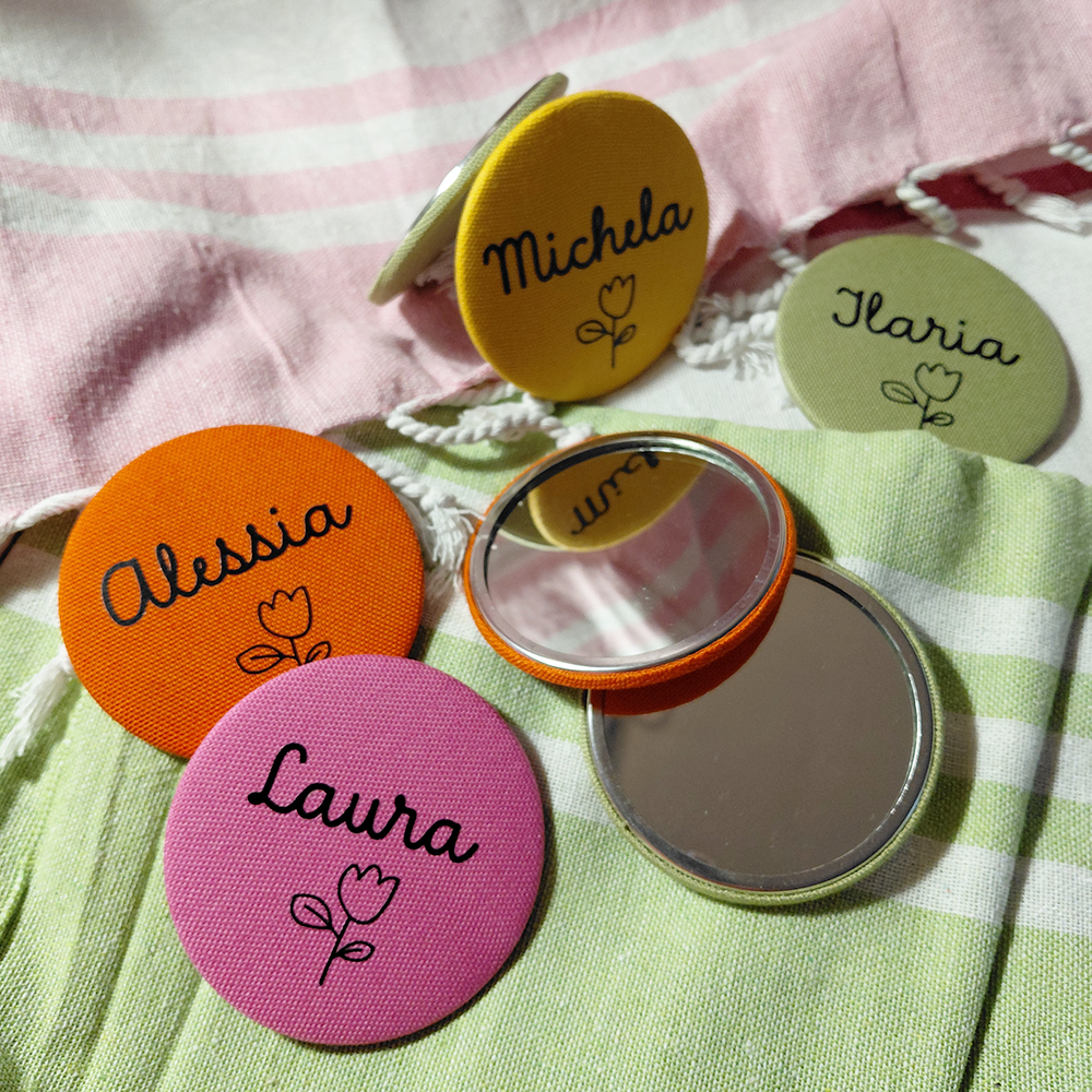 Quattro badge colorati e due "Specchietto tondo colorato e personalizzato" di In Casa di Ale poggiano su un tessuto a righe pastello.