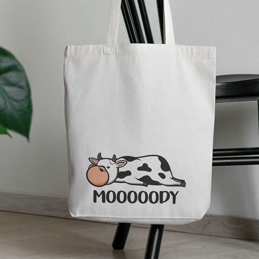 Una Tote bag con illustrazioni di Il mio negozio in cotone bianco è appesa a una sedia nera. Presenta una mucca brontolona e la scritta "MOOOOODY" in basso. Una pianta fa capolino sulla sinistra: un regalo originale per ogni stato d'animo!.